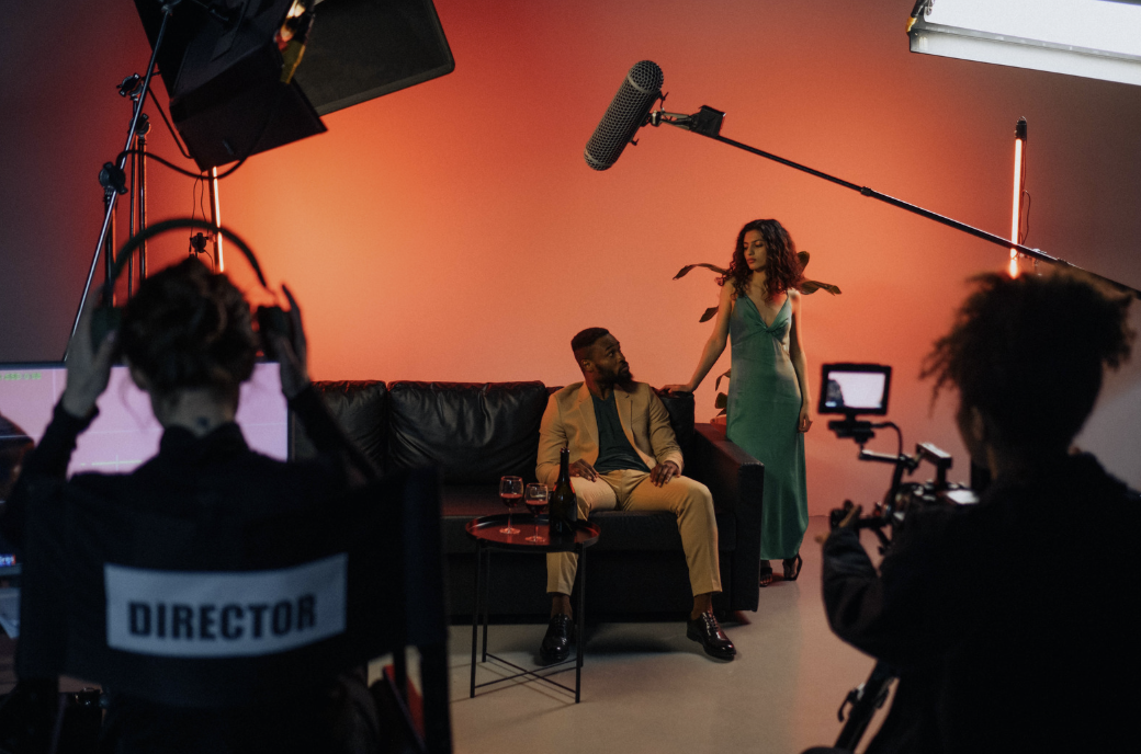 Two actors stand on set with cameras, lights, and a boom mic around them.